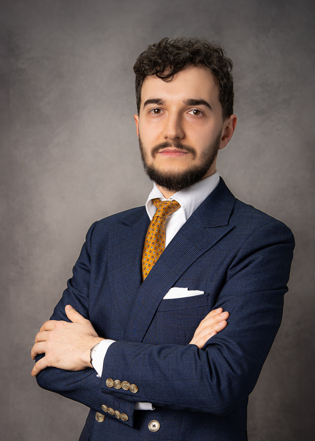 Portret młodego adwokata w granatowym garniturze w kratę i żółtym krawacie, z rękami skrzyżowanymi na piersi, na tle szarego tła. Profesjonalny wizerunek.