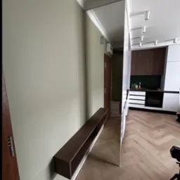 Nowoczesny salon z minimalistyczną szafką RTV zawieszoną na ścianie i lustrem. Widoczna kuchnia w tle, parkiet w jodełkę, laserowy poziom na ścianie.