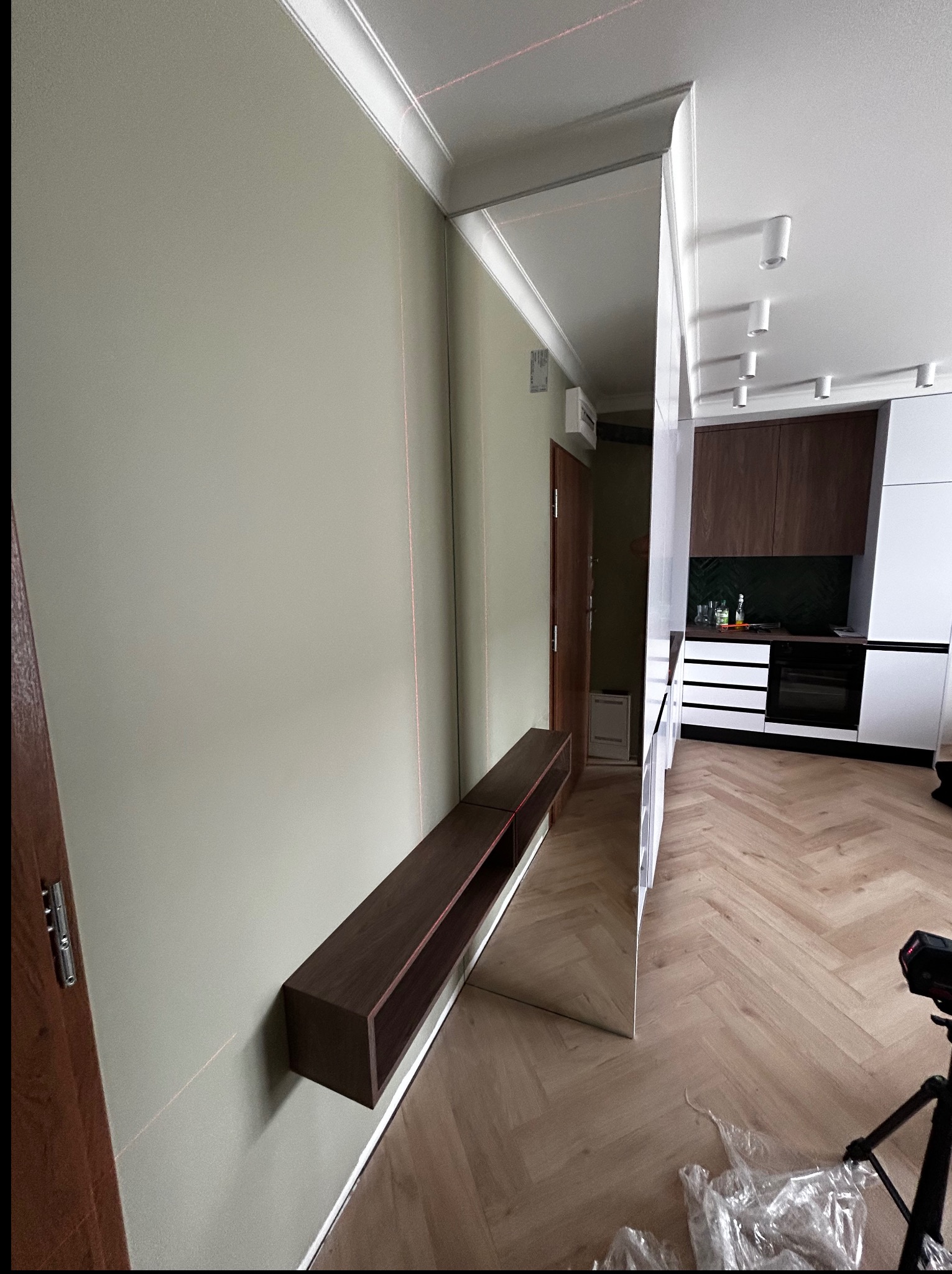 Nowoczesny salon z minimalistyczną szafką RTV zawieszoną na ścianie i lustrem. Widoczna kuchnia w tle, parkiet w jodełkę, laserowy poziom na ścianie.