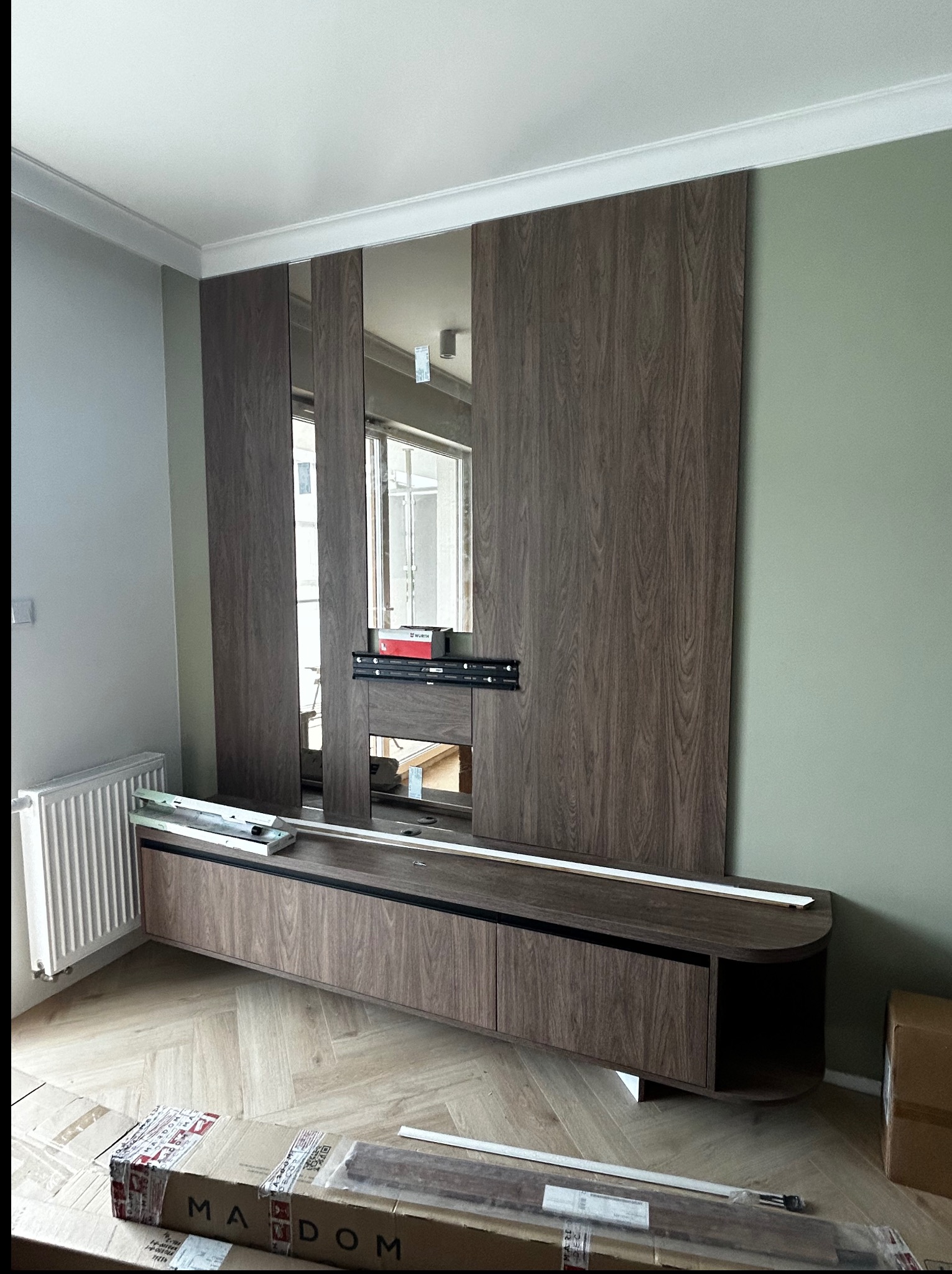 Drewniana zabudowa TV z lustrem w salonie, w trakcie montażu. Widoczne elementy montażowe i kartony na podłodze. Parkiet w jodełkę, ściany w odcieniach szarości i zieleni.