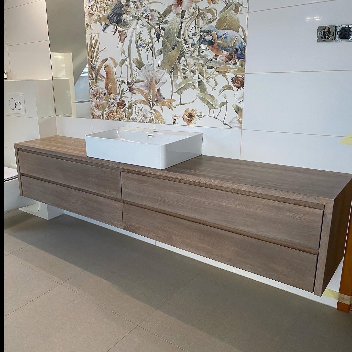 Nowoczesna, wisząca szafka łazienkowa z drewna w odcieniach brązu, z białą umywalką nablatową. W tle białe płytki z motywem roślinnym i lustro.