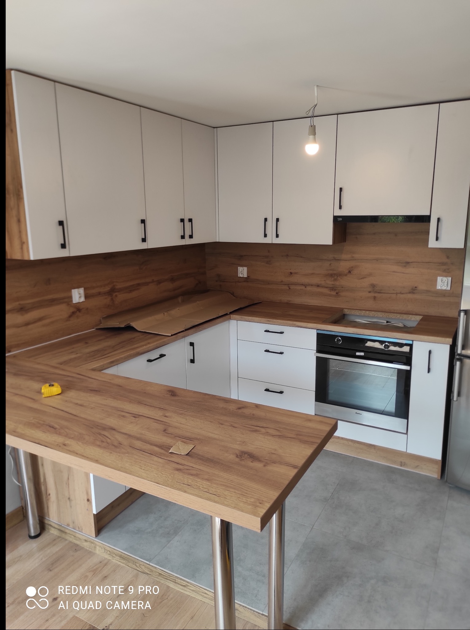 Jasna kuchnia z drewnianym blatem i barkiem na metalowych nogach. Widoczny fragment kartonu na blacie, sugerujący prace wykończeniowe. Nowoczesne fronty.