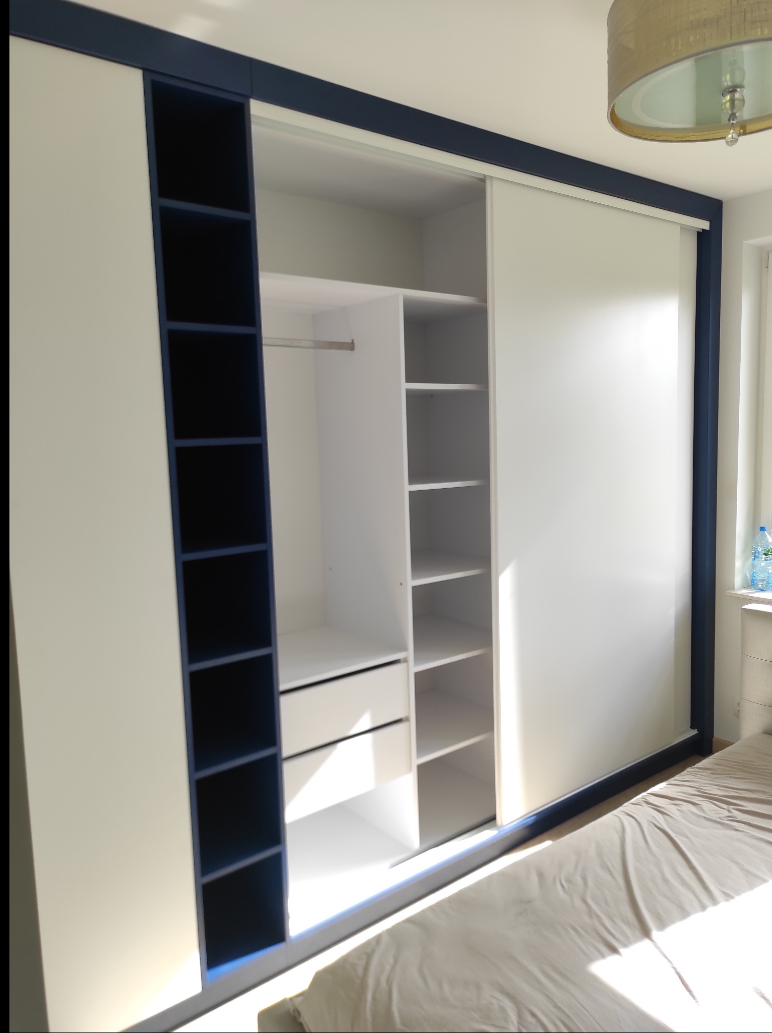 Biała szafa wnękowa z przesuwnymi drzwiami i niebieskimi akcentami, widoczna w sypialni, z otwartymi półkami i szufladami, oświetlona naturalnym światłem.