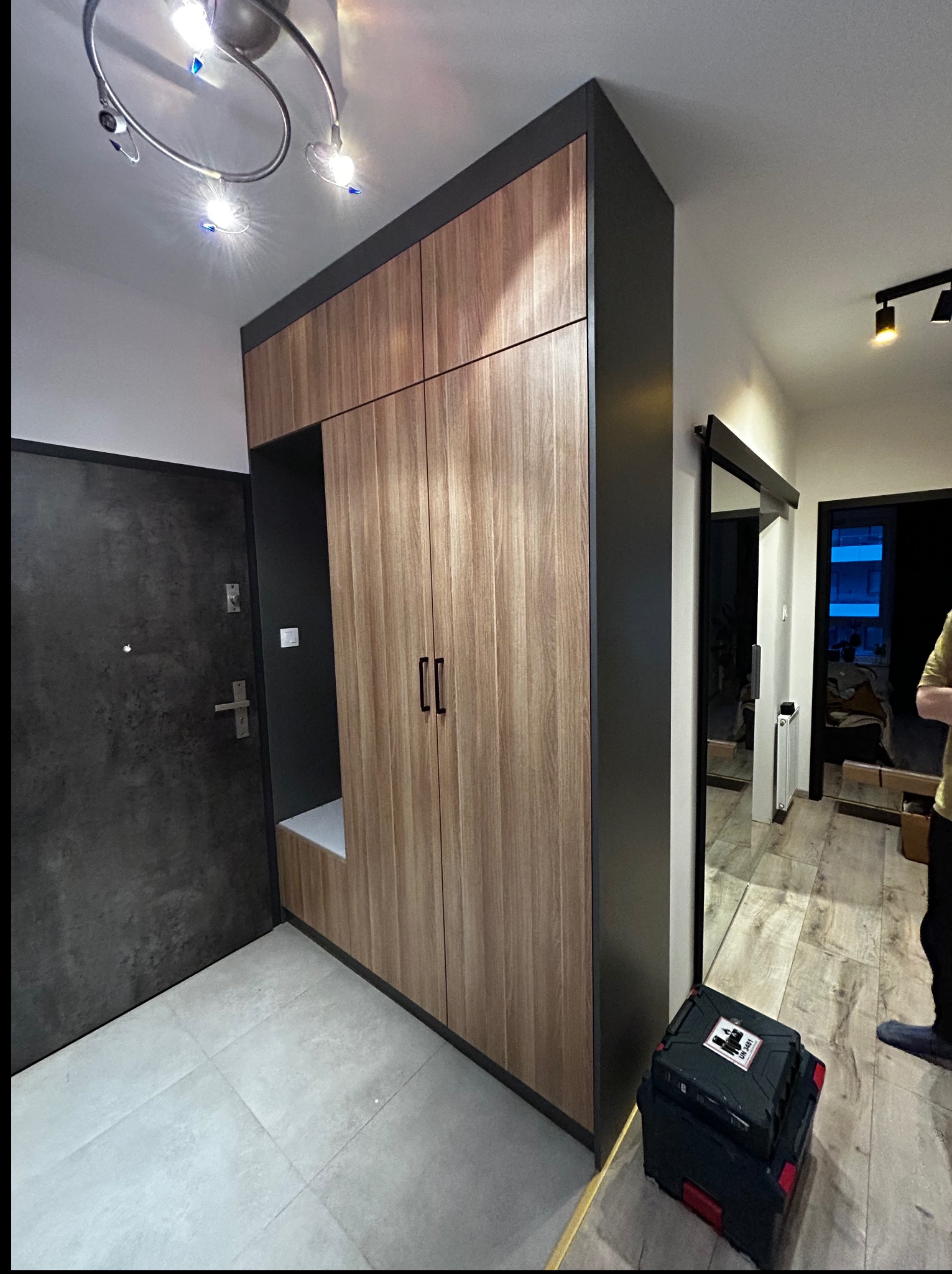 Funkcjonalna szafa wnękowa z siedziskiem w przedpokoju, wykończona drewnopodobną okleiną i antracytowymi detalami. Widoczna skrzynka narzędziowa sugeruje świeży montaż.