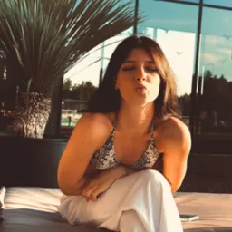 Kobieta w bikini z motywem skóry węża, siedząca na sofie na tle nowoczesnego budynku z palmą. Usta ułożone w dzióbek, oczy zamknięte. Ciepłe światło słoneczne.