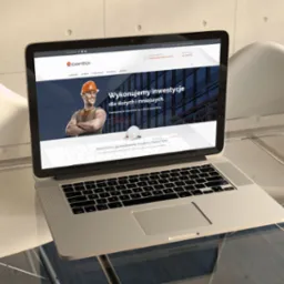 Laptop na szklanym stole prezentuje stronę internetową firmy budowlanej z hasłem 'Wykonujemy inwestycje dla dużych i mniejszych'. W tle nowoczesne krzesła i betonowa ściana.