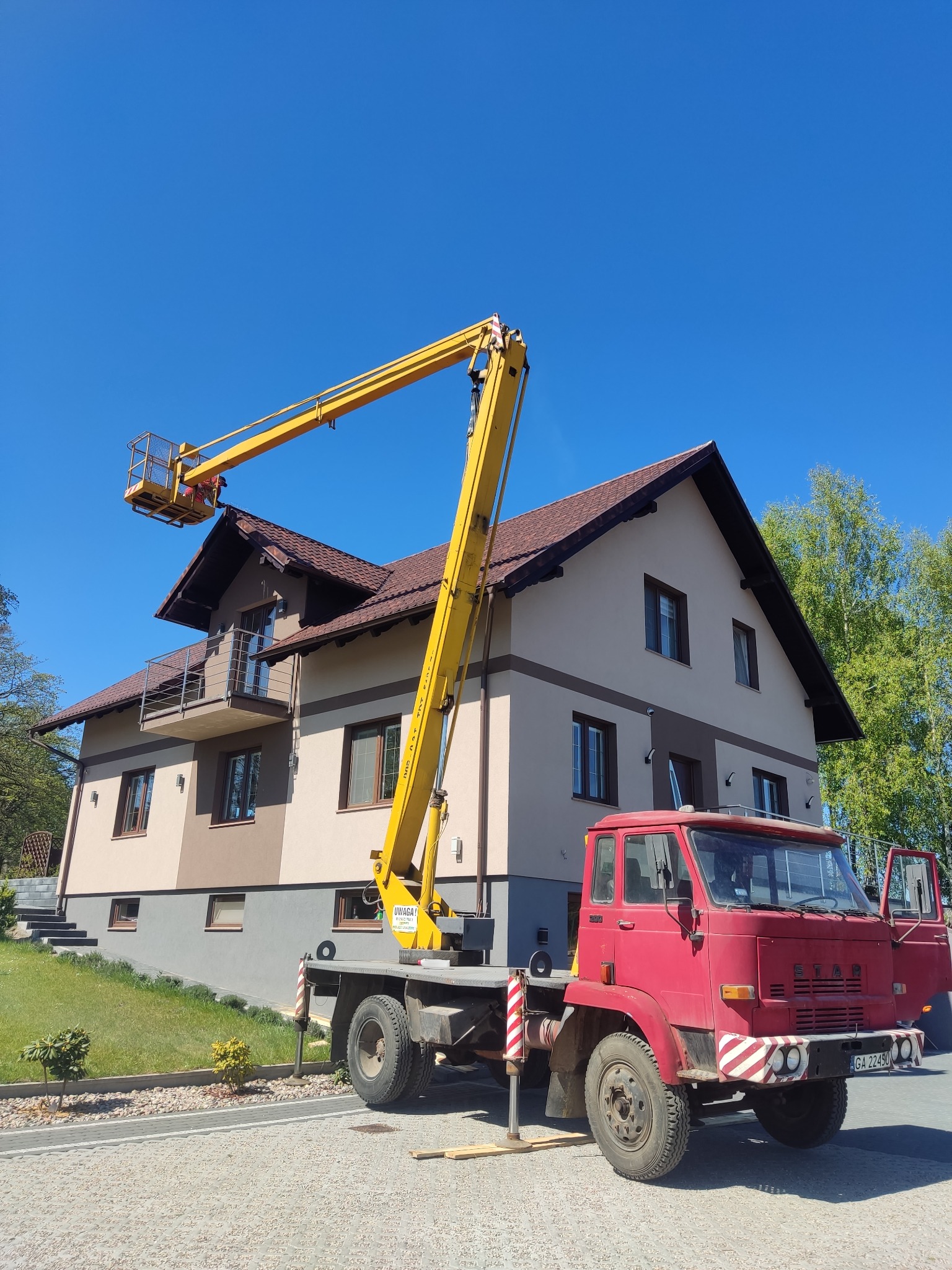 Czerwony podnośnik koszowy z żółtym ramieniem na tle dwukondygnacyjnego domu z brązowym dachem i błękitnego nieba. Widoczny fragment zielonego trawnika i drzew.