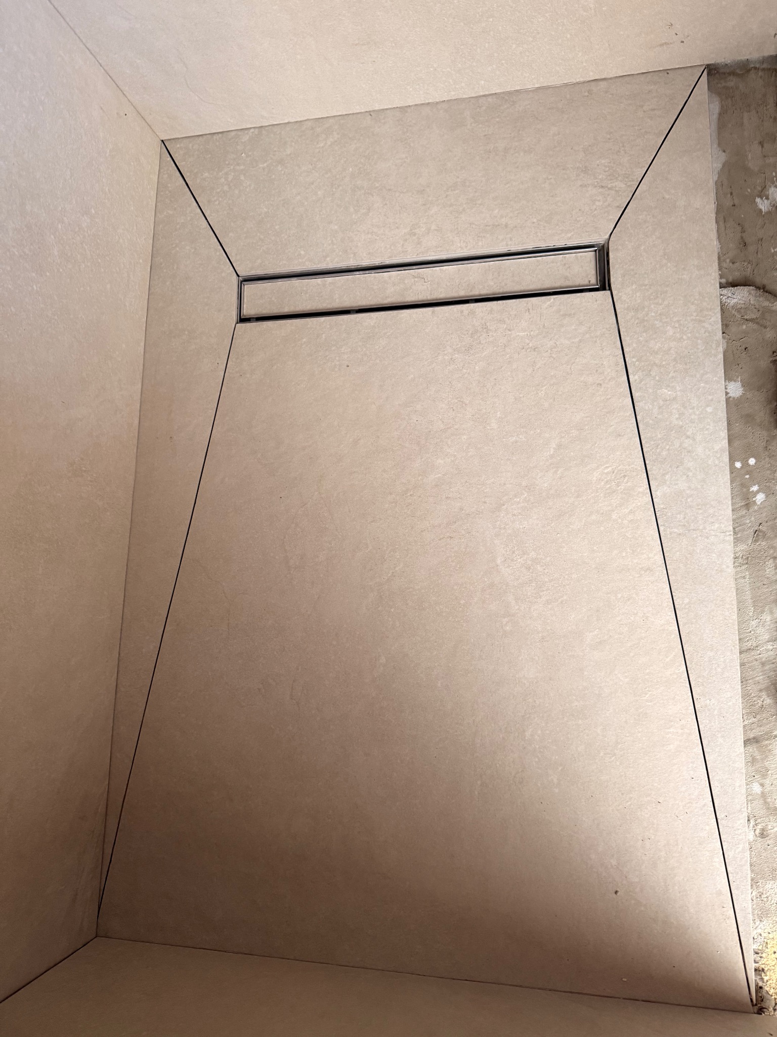 Wykończony brodzik z odpływem liniowym, obłożony beżowymi płytkami imitującymi kamień. Widoczne fugi i fragment ściany przed wykończeniem.