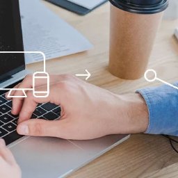 Vertes Design - strony internetowe Warszawa - Mężczyzna pisze na laptopie z grafiką booking i ikoną komputera/telefonu. Widoczne dłonie, fragment twarzy, kawa na biurku i notatki. Jasne, naturalne światło.
