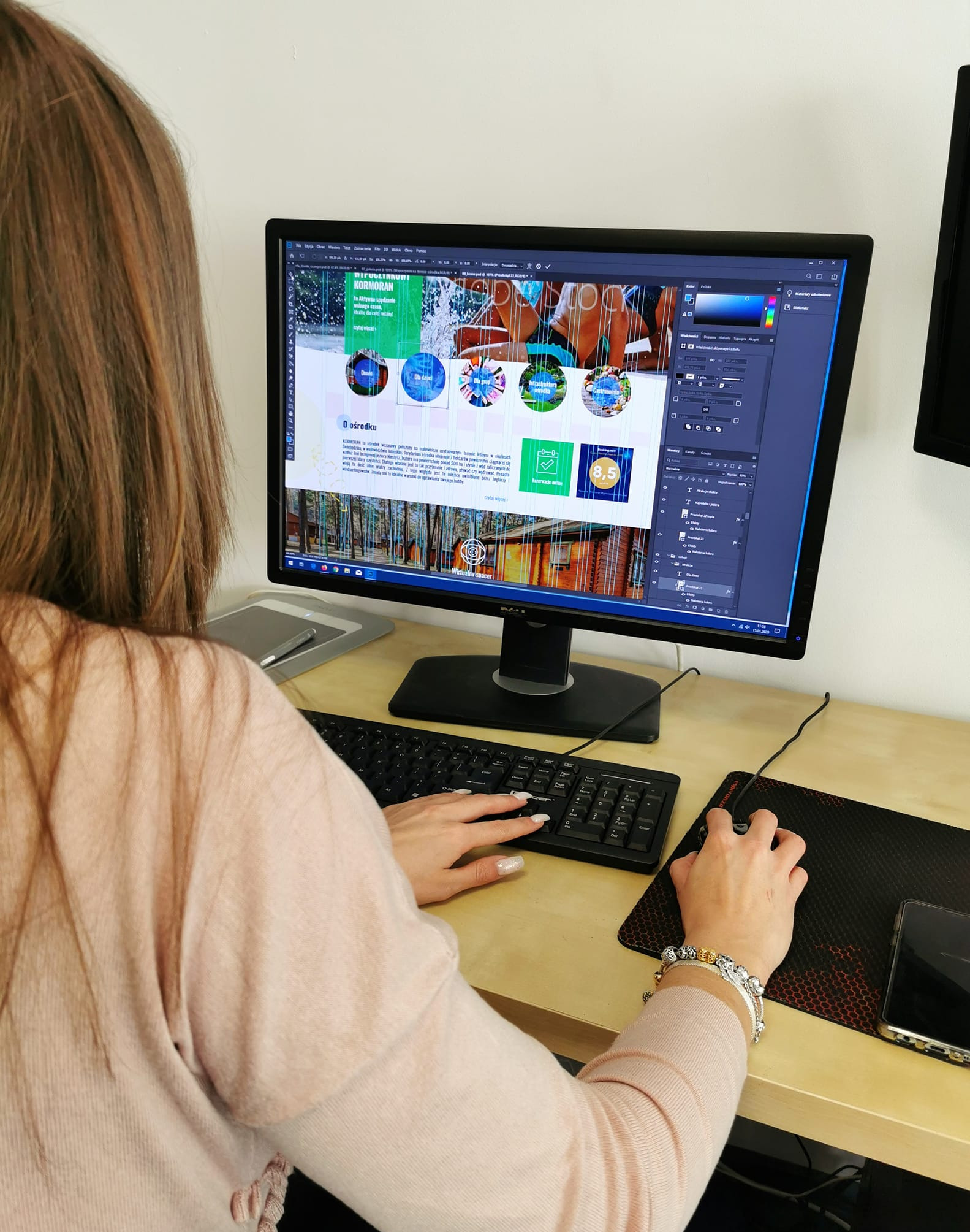 Graficzka pracuje nad projektem strony internetowej ośrodka wypoczynkowego, używając programu graficznego na monitorze. Widoczna klawiatura, myszka i tablet graficzny.