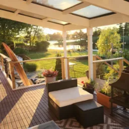 Zadaszony taras z drewnianą podłogą i widokiem na jezioro o zachodzie słońca. Elementy dekoracyjne: deska surfingowa, palenisko, rattanowe meble.