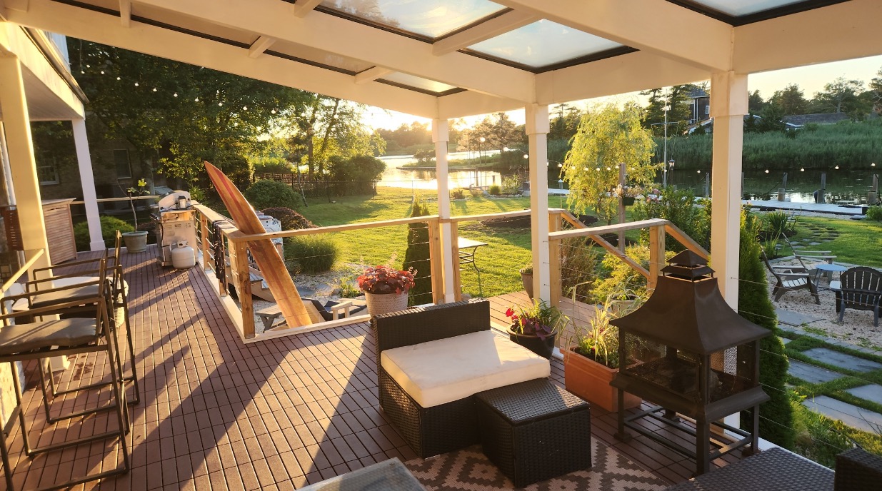 Zadaszony taras z drewnianą podłogą i widokiem na jezioro o zachodzie słońca. Elementy dekoracyjne: deska surfingowa, palenisko, rattanowe meble.