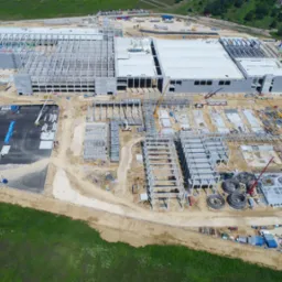 Widok z lotu ptaka na plac budowy hali przemysłowej w trakcie wznoszenia konstrukcji stalowej i betonowej. Widoczne dźwigi i maszyny budowlane.