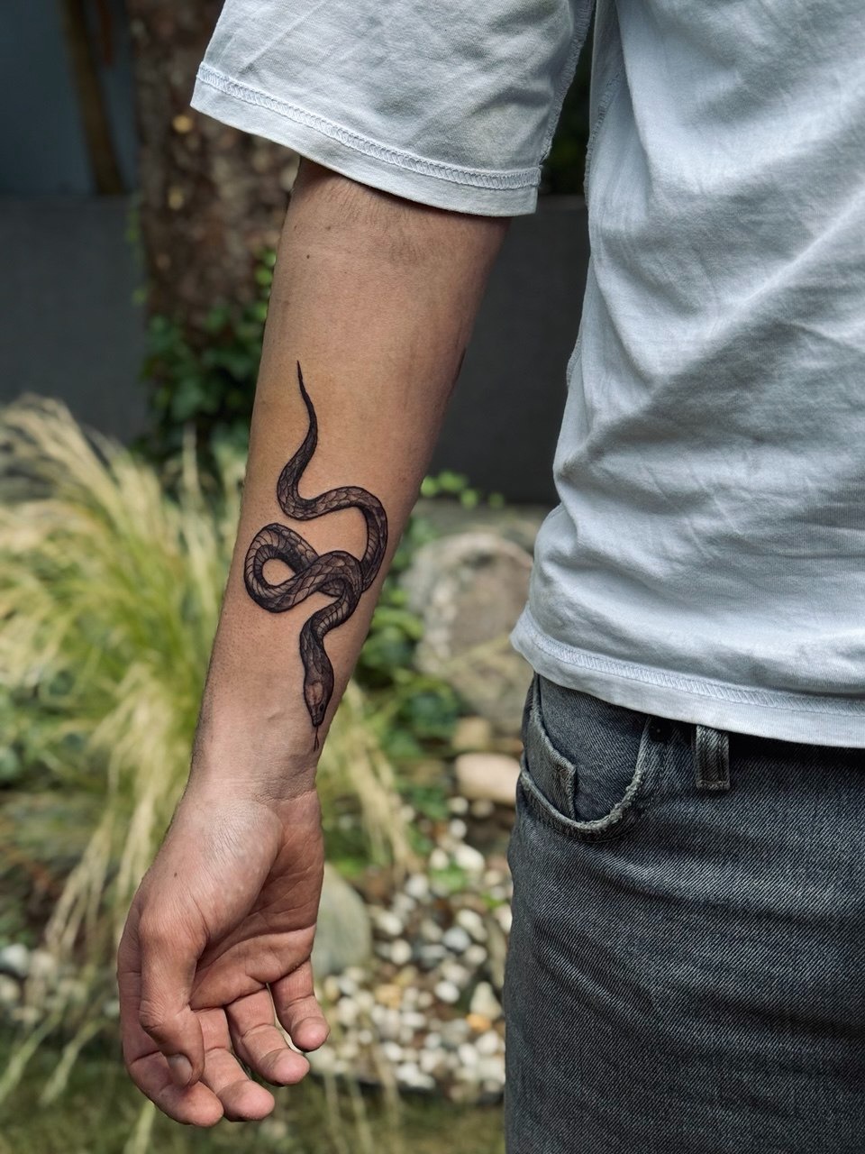 Ramię z tatuażem czarnej kobry na tle ogrodu. Widoczne detale wzoru i cieniowania. Realistyczny styl tatuażu.