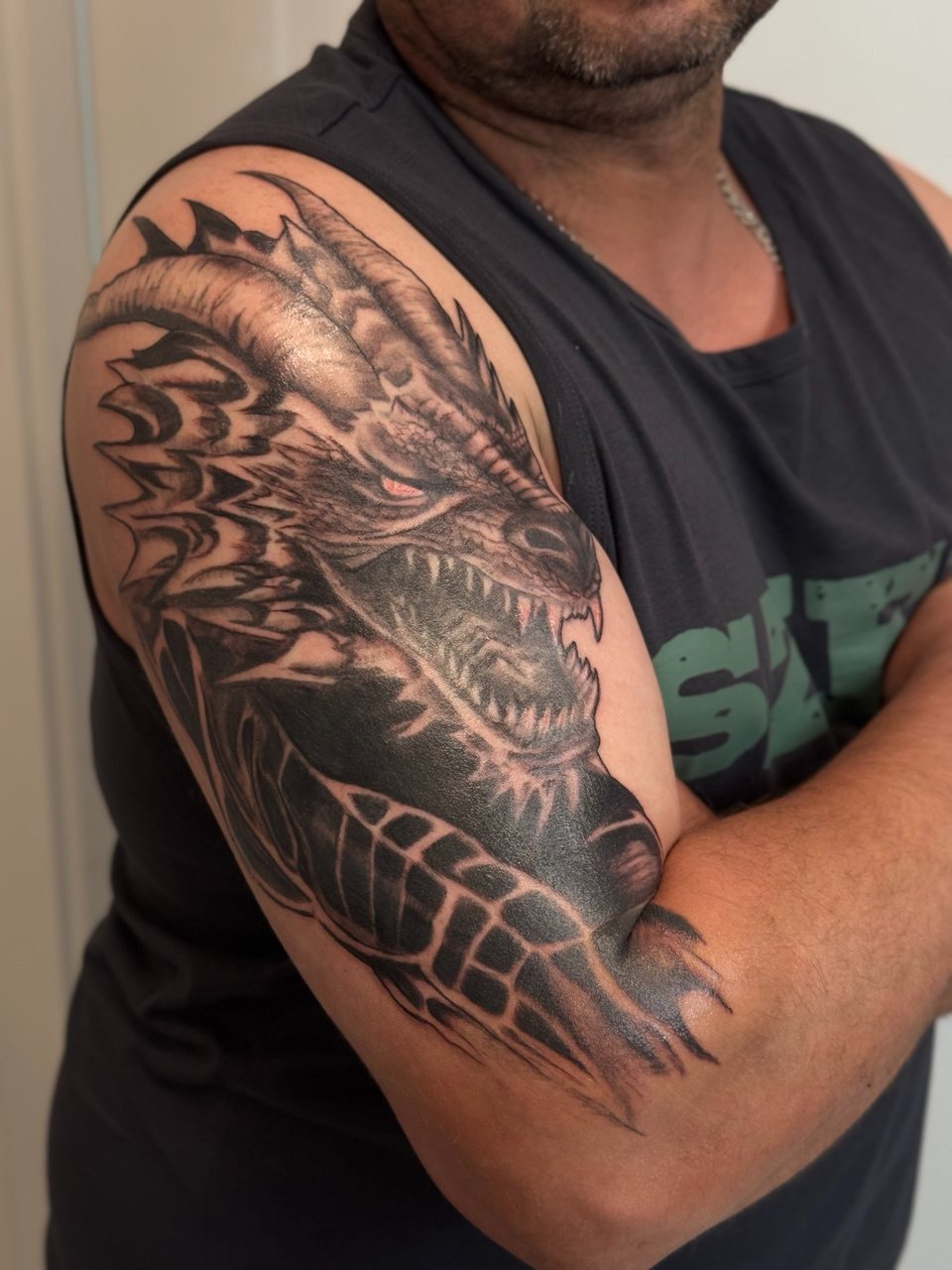 Ramię mężczyzny z czarno-szarym tatuażem smoka z czerwonymi oczami, widoczne zęby i rogi, na tle jasnej ściany. Tatuaż obejmuje biceps i część przedramienia.