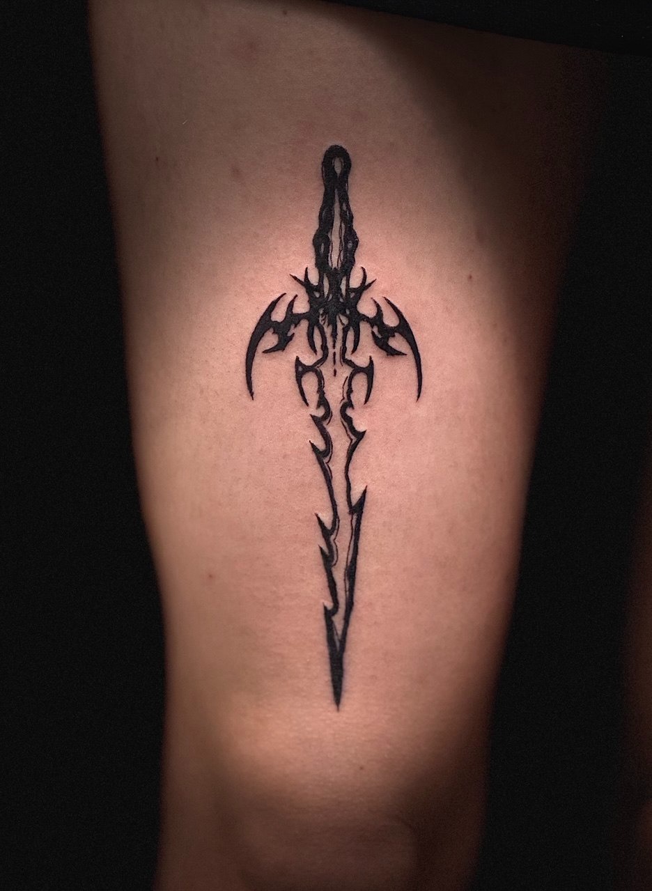 Czarny, gotycki tatuaż miecza na jasnej skórze, umieszczony na udzie, z widocznymi drobnymi niedoskonałościami linii. Ciemne tło podkreśla wzór.