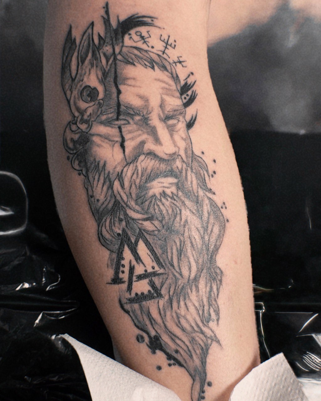 Tatuaż na ramieniu przedstawiający portret brodatego mężczyzny z motywami mitologicznymi, wykonany w czerni i szarości, z dbałością o detale cieniowania.