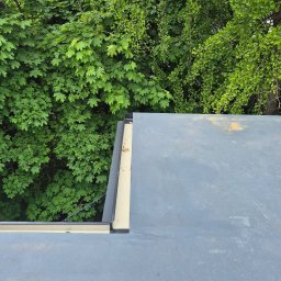 Len-dach - Szary dach z widoczną obróbką blacharską, na tle zielonych drzew. Ujęcie z góry prezentuje detale pokrycia i estetyczne wykończenie dachu.