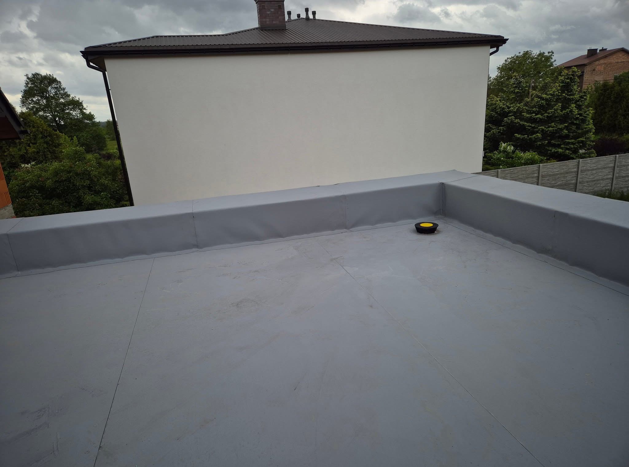 Szary, płaski dach pokryty papą termozgrzewalną z żółtym odpowietrznikiem. W tle budynek z brązowym dachem i kominem oraz drzewa pod pochmurnym niebem.