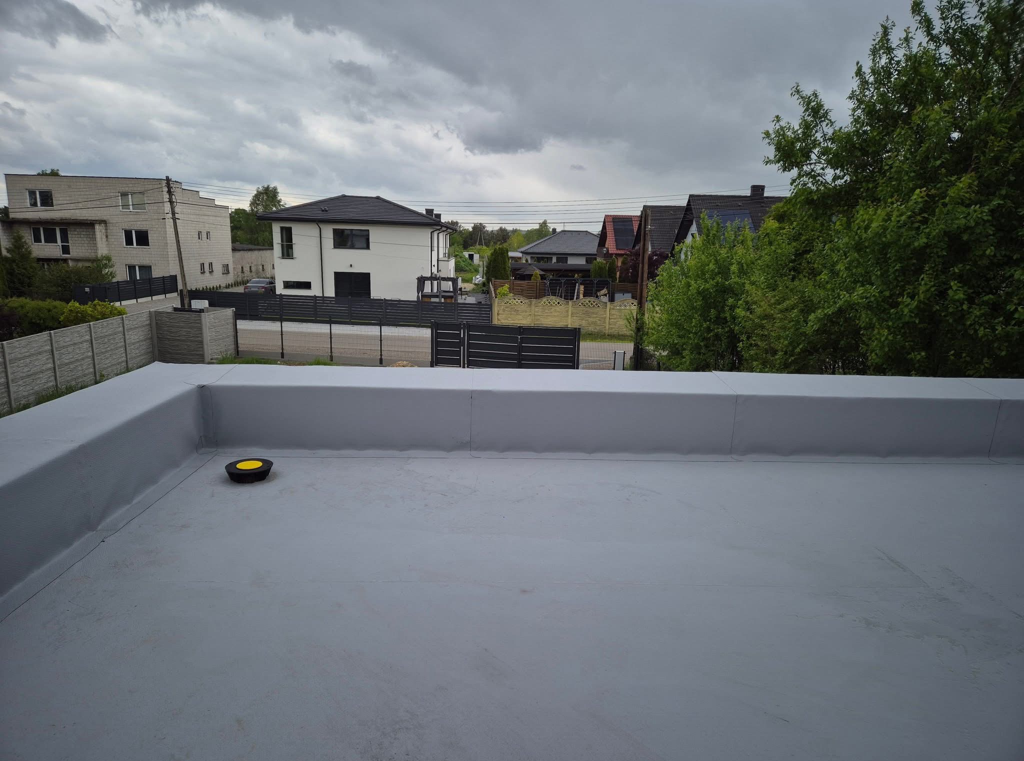 Szary, płaski dach z widocznymi łączeniami i kominkiem wentylacyjnym na tle zabudowy jednorodzinnej pod pochmurnym niebem.