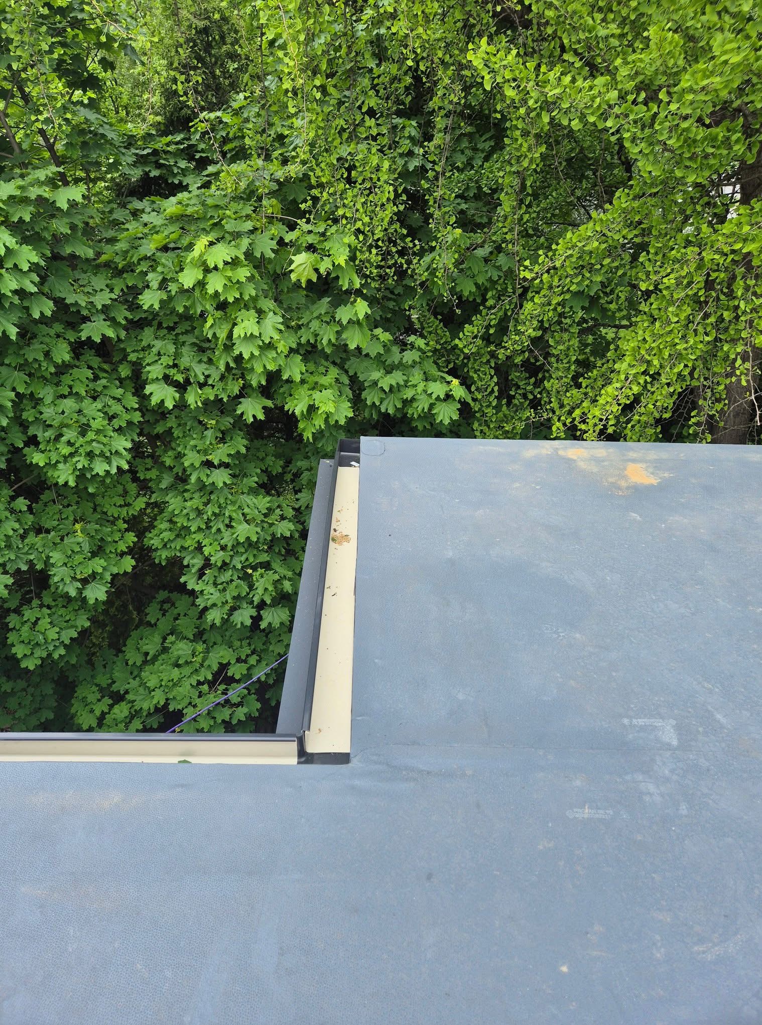 Szary dach z widoczną obróbką blacharską, na tle zielonych drzew. Ujęcie z góry prezentuje detale pokrycia i estetyczne wykończenie dachu.