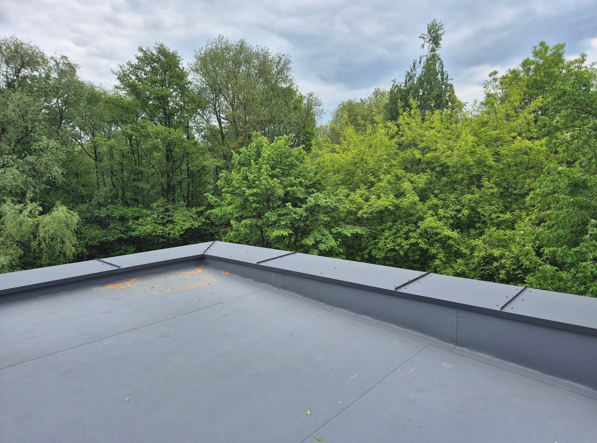 Szary, płaski dach z obróbkami blacharskimi na tle zieleni drzew i pochmurnego nieba. Widok z góry.