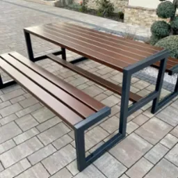 Stół piknikowy z ławkami o nowoczesnej, minimalistycznej konstrukcji z ciemnego metalu i drewnianymi siedziskami, ustawiony na kostce brukowej.