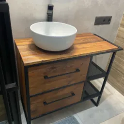 Loftowa szafka łazienkowa z drewnianymi szufladami i metalową konstrukcją, na blacie umywalka nablatowa. Widoczne gniazdko elektryczne w tle.