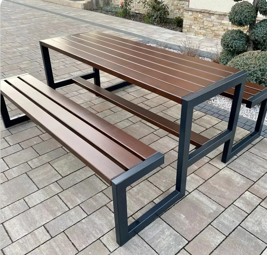 Stół piknikowy z ławkami o nowoczesnej, minimalistycznej konstrukcji z ciemnego metalu i drewnianymi siedziskami, ustawiony na kostce brukowej.