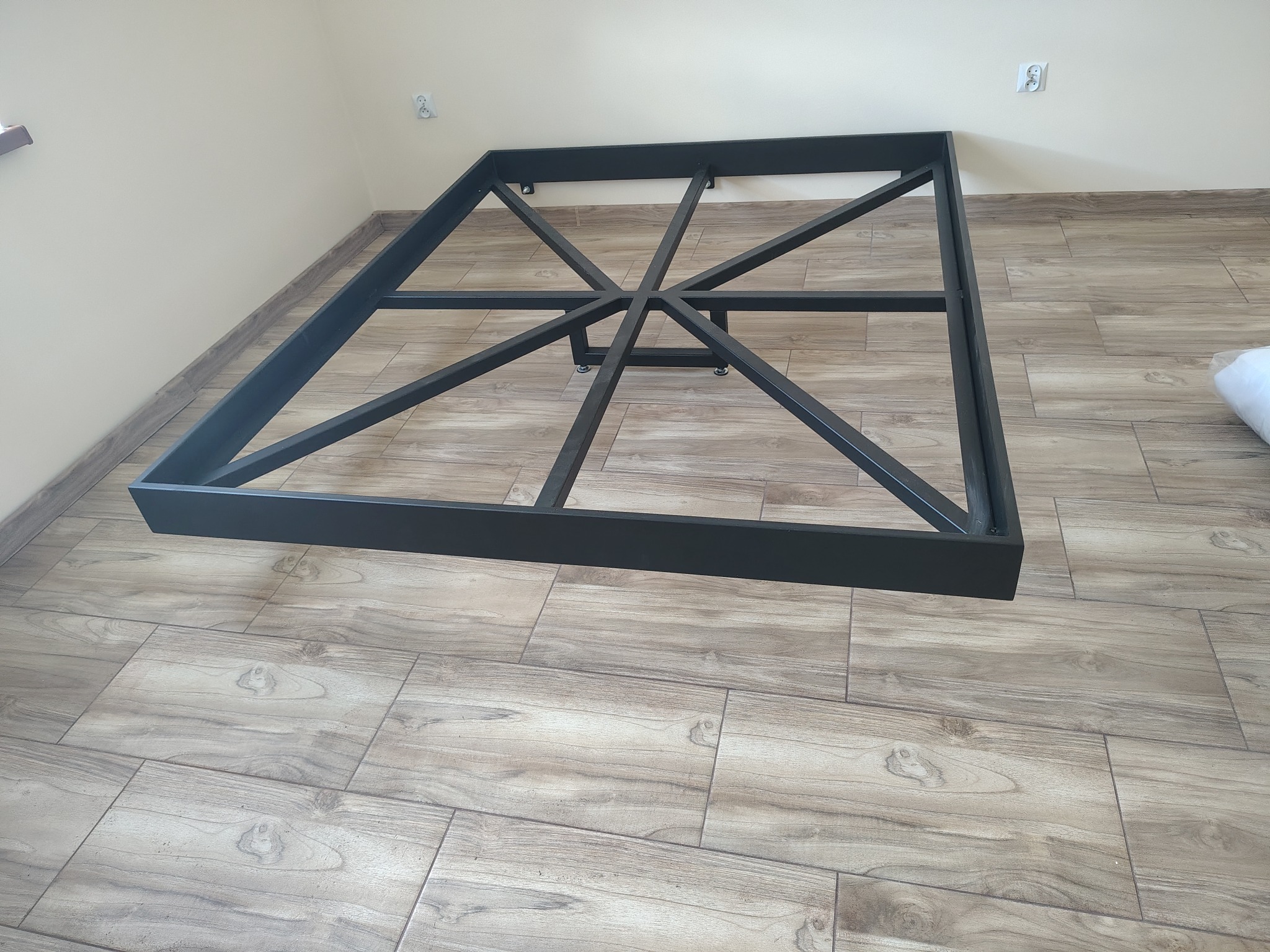 Czarna, metalowa konstrukcja ramy łóżka z centralnym wsparciem, ustawiona na podłodze z płytek imitujących drewno, widok z perspektywy, jasne ściany w tle.