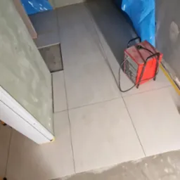 Układanie jasnych, kwadratowych płytek podłogowych w łazience w trakcie remontu. Widoczna nagrzewnica i narzędzia glazurnicze.