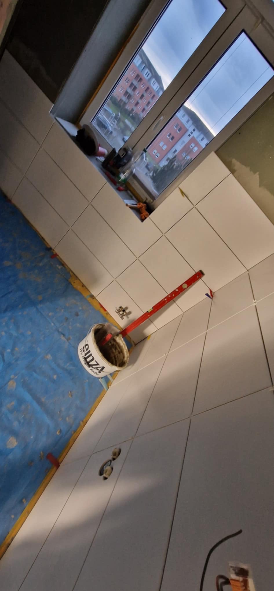 Białe płytki ułożone na ścianie w łazience, widoczna poziomica i wiadro z klejem, okno w tle. Praca w toku, etap układania płytek.