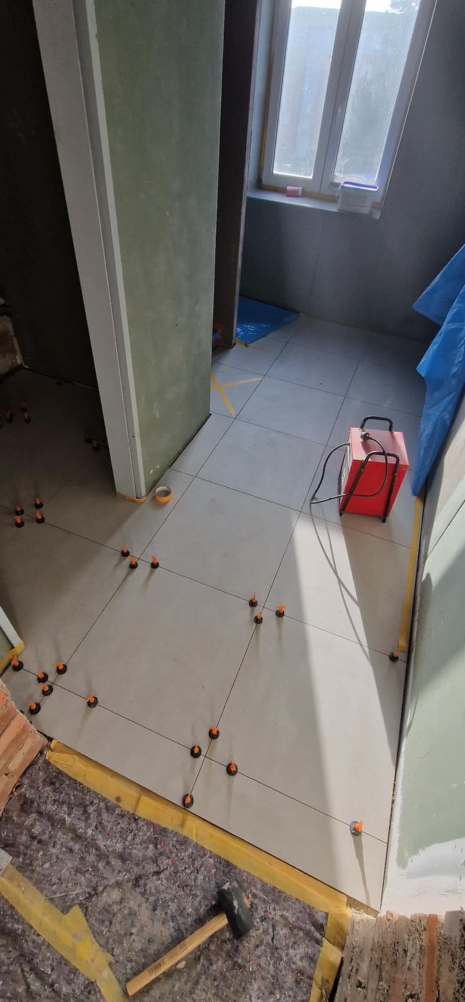 Ułożone jasne płytki podłogowe z systemem poziomowania, widoczny fragment podłoża z młotkiem, w tle okno i grzejnik. Etap prac glazurniczych.