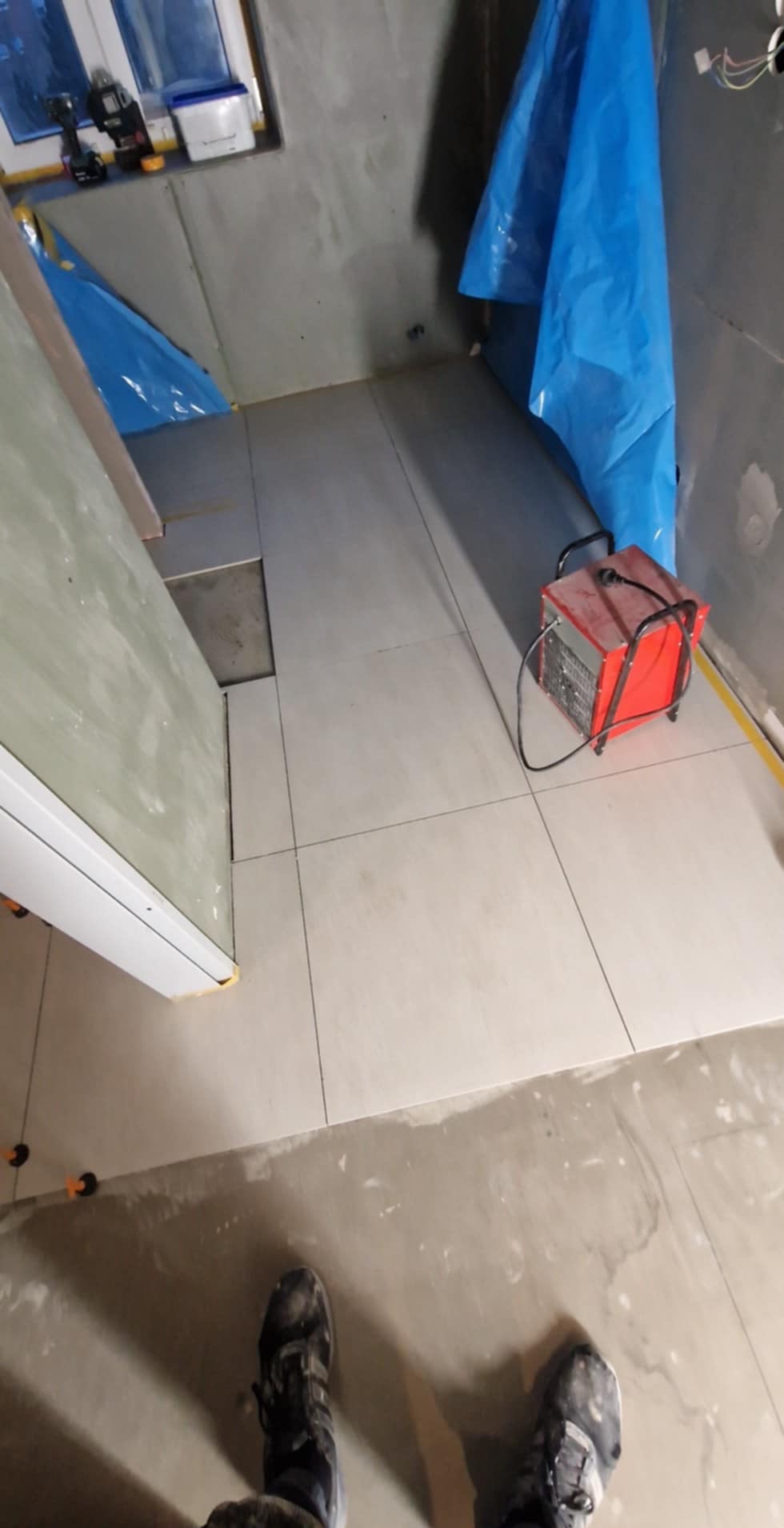 Układanie jasnych, kwadratowych płytek podłogowych w łazience w trakcie remontu. Widoczna nagrzewnica i narzędzia glazurnicze.