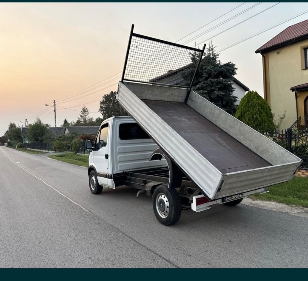 Biały samochód dostawczy z otwartą skrzynią ładunkową na ulicy, widok z tyłu, w tle domy jednorodzinne i zachodzące słońce.