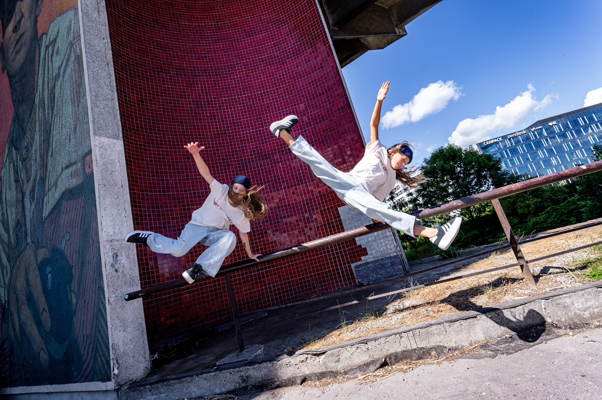 Dwie młode kobiety w dynamicznych pozach tanecznych na tle miejskiego krajobrazu z mozaiką i nowoczesnym budynkiem w tle, podkreślone intensywnym światłem słonecznym.