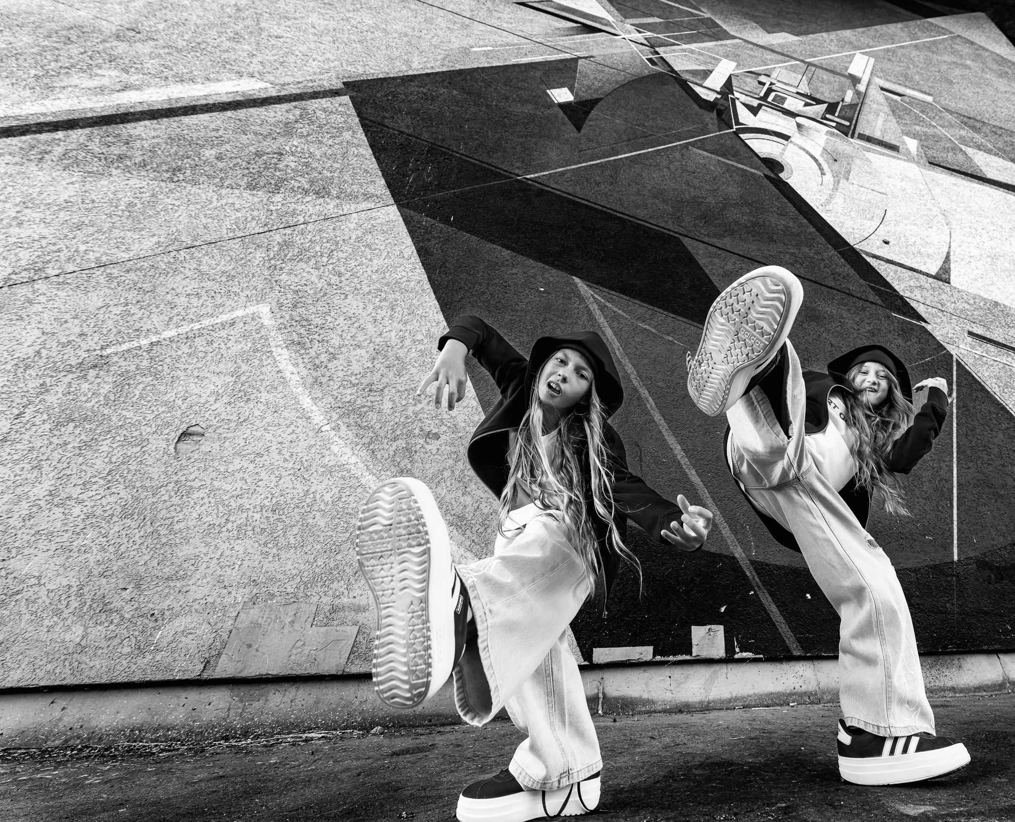 Dwie dziewczyny w szerokich spodniach i butach Adidas, pozują w tanecznych ruchach na tle abstrakcyjnego muralu w czerni i bieli. Dynamiczne ujęcie z dołu.