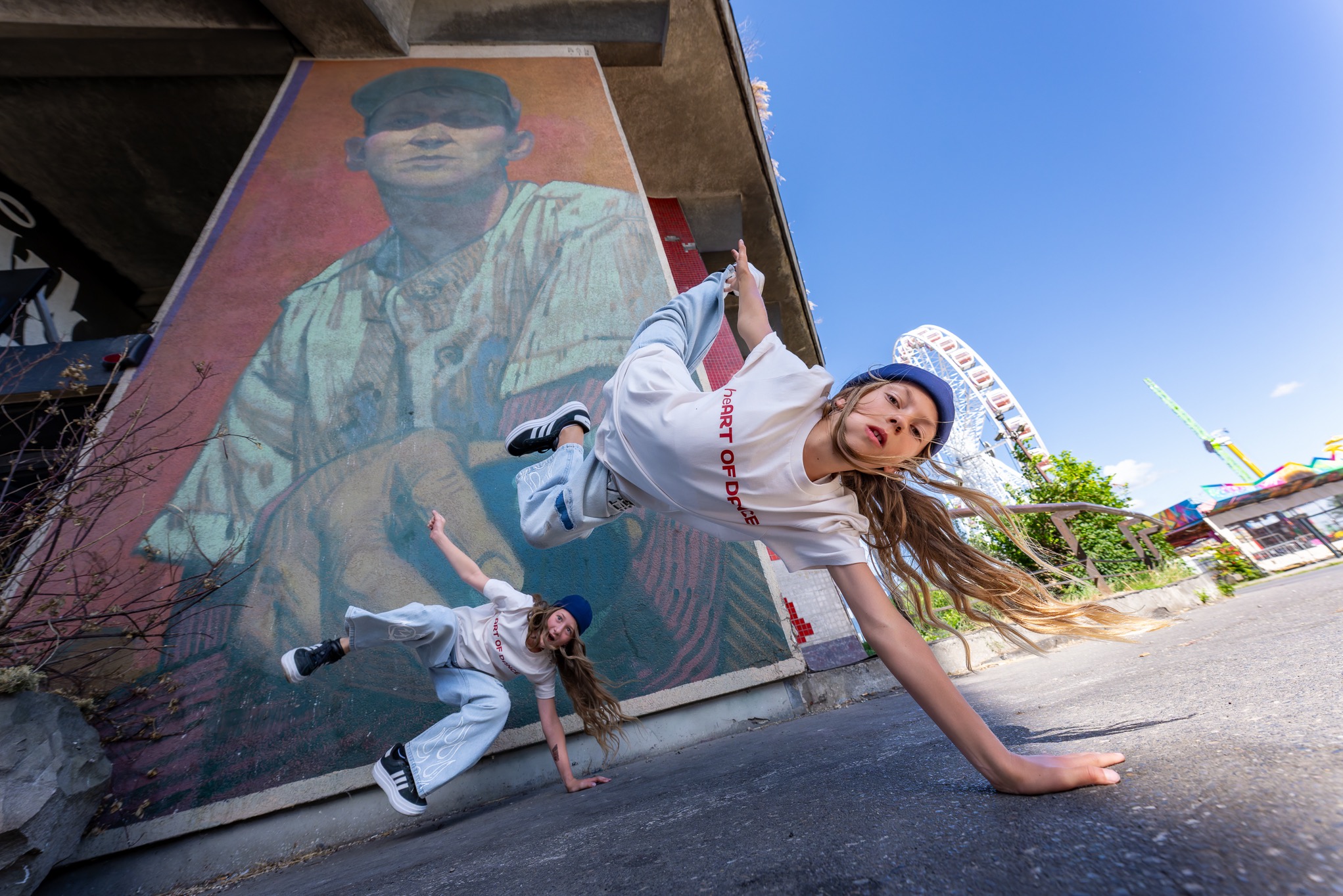 Dwie młode tancerki w dynamicznych pozach na tle muralu i wesołego miasteczka. Dziewczyny w identycznych strojach prezentują umiejętności taneczne.