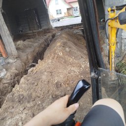 Rem-Bud - Z perspektywy operatora: Żółta koparka w trakcie wykopu wąskiego rowu pod fundamenty budynku. W tle widoczny dom i przyczepa.