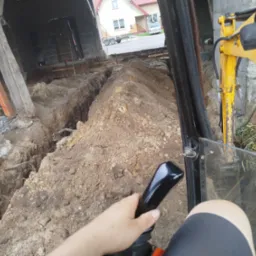 Z perspektywy operatora: Żółta koparka w trakcie wykopu wąskiego rowu pod fundamenty budynku. W tle widoczny dom i przyczepa.
