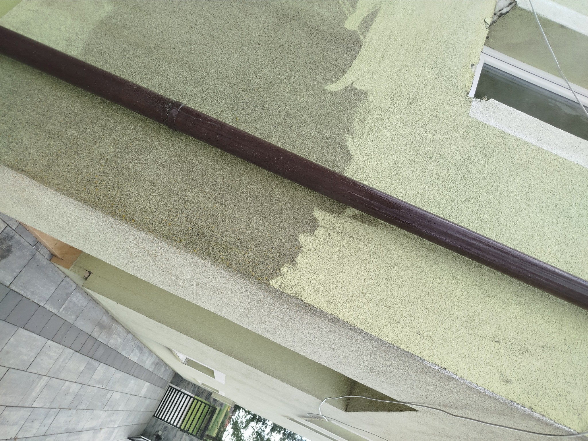 Fragment elewacji budynku z rynną i świeżo pomalowaną, jasną elewacją, widoczne ślady po malowaniu. Kąt ujęcia z dołu, widoczny fragment chodnika.