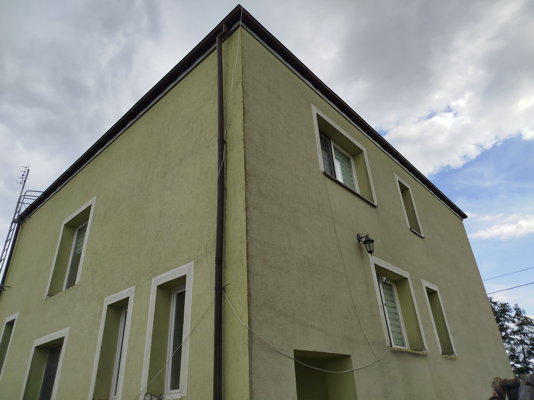 Dwukondygnacyjny budynek z oknami w białych ramach, otynkowany na zielono, widziany z dołu pod kątem, z widocznym fragmentem dachu i niebem z chmurami.