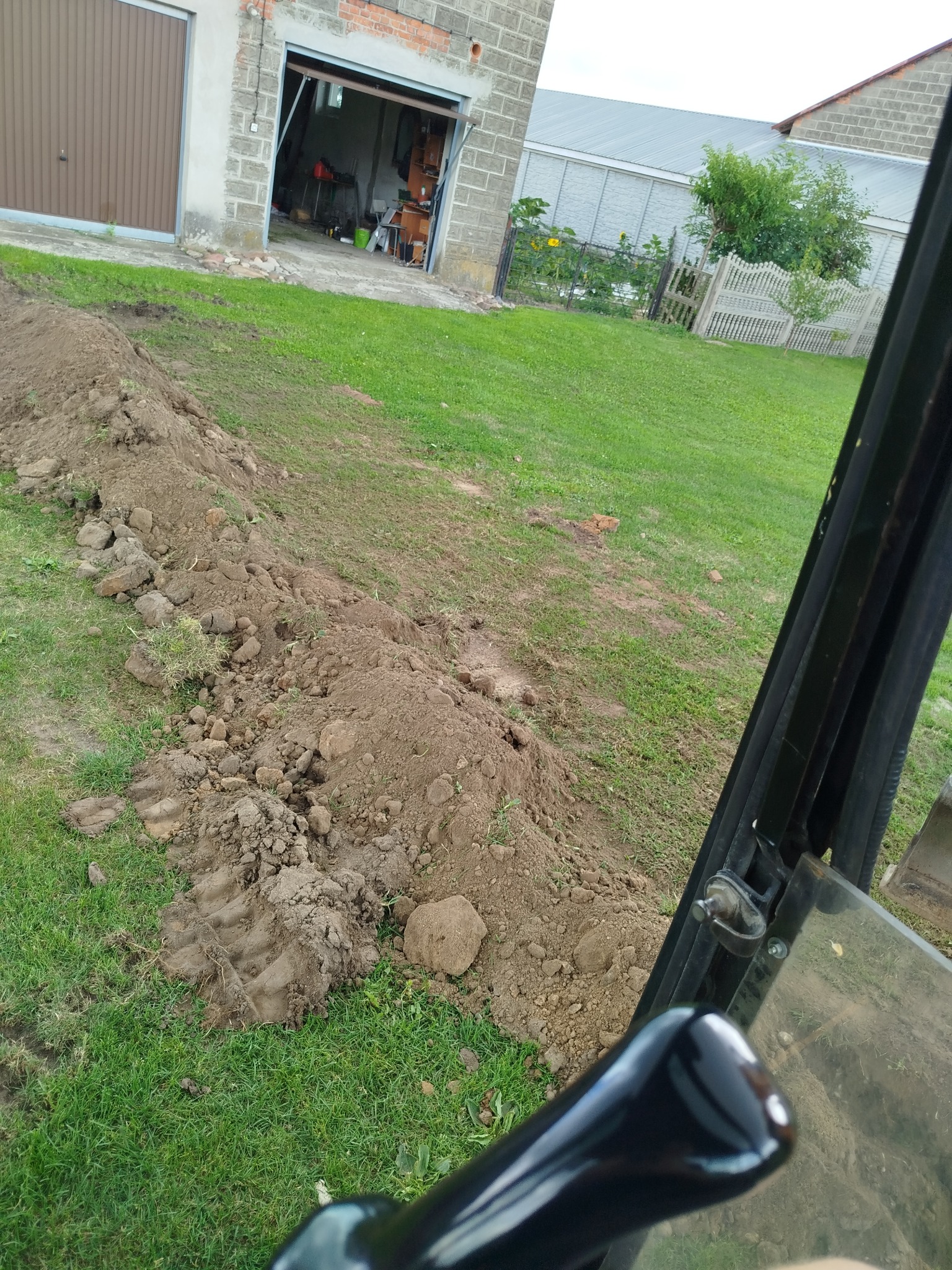 Świeżo wykopany rów na trawiastym terenie, widok z kabiny maszyny budowlanej. W tle budynek gospodarczy i zieleń.