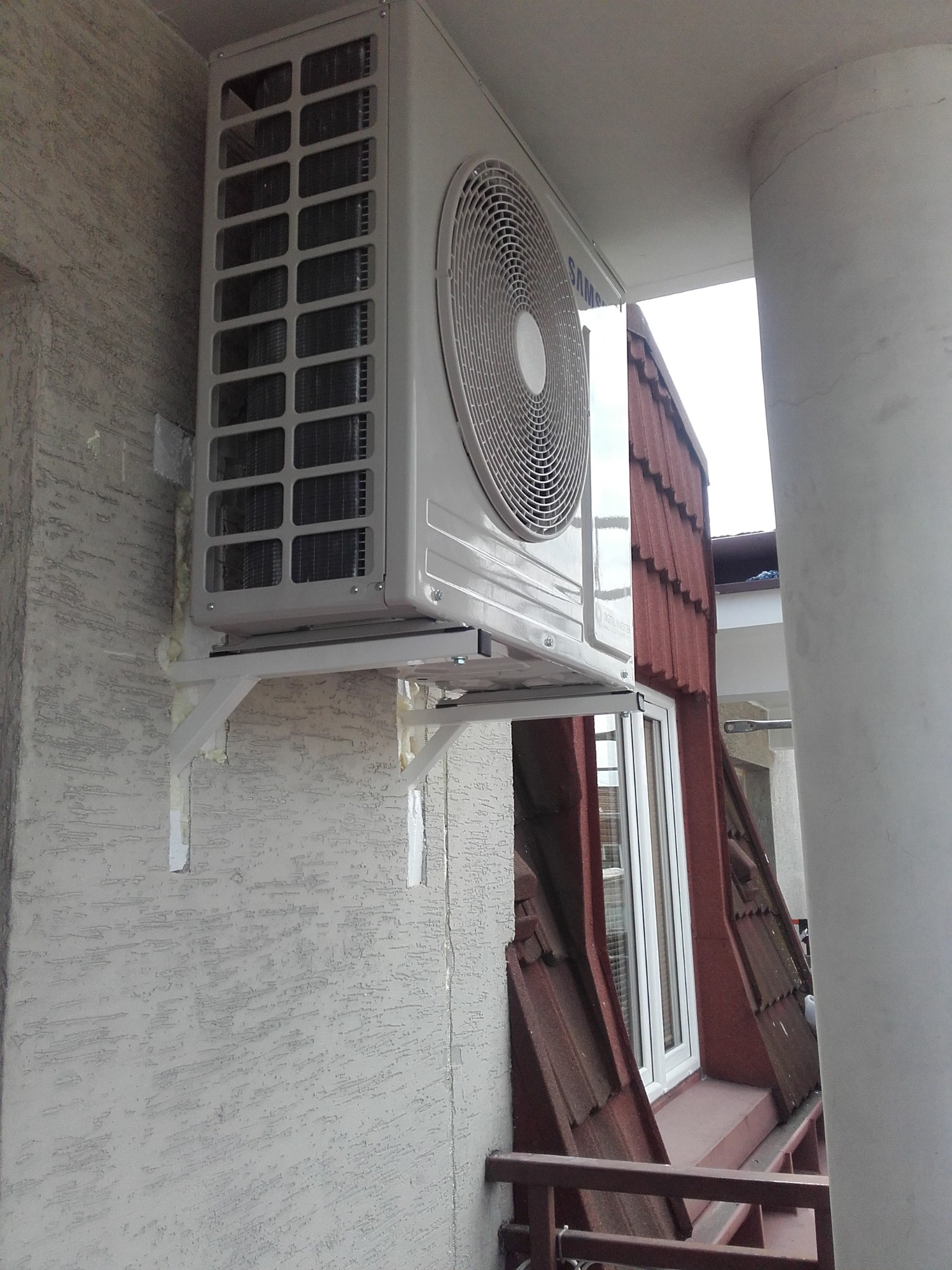 Zamontowana klimatyzacja Samsung na zewnątrz budynku, widoczna na wsporniku. Biała obudowa kontrastuje z szarą elewacją i fragmentem dachu z czerwonej dachówki.