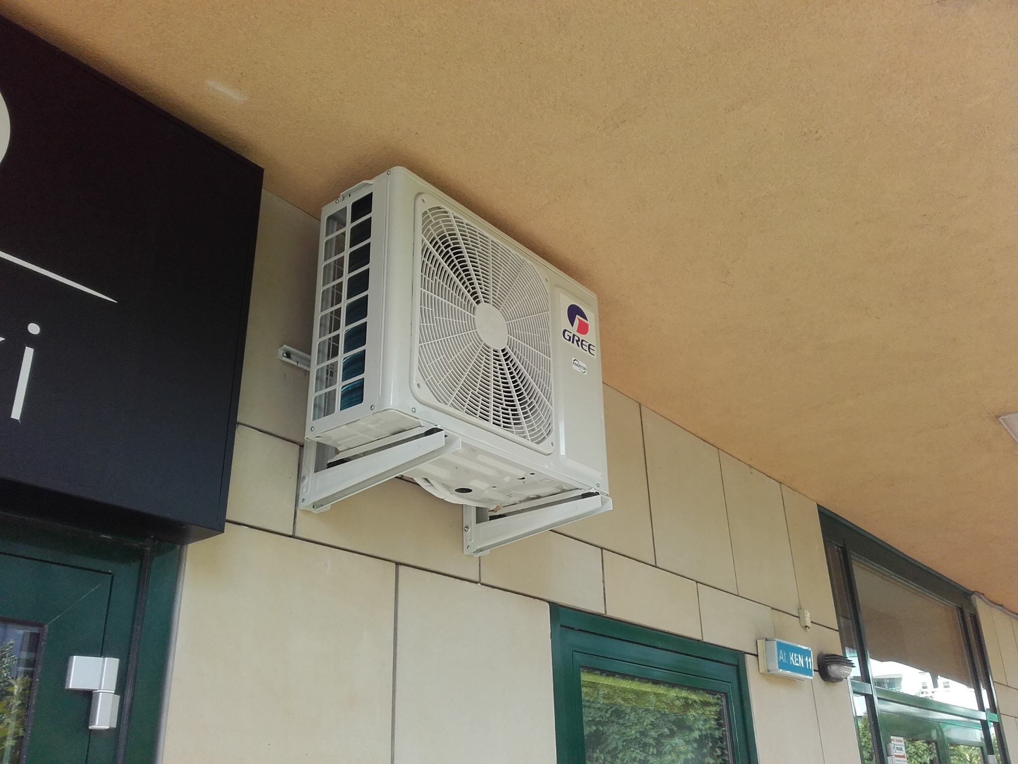 Biały klimatyzator marki Gree zamontowany na elewacji budynku z beżowymi płytkami. Widoczny fragment szyldu i okna z zieloną ramą.