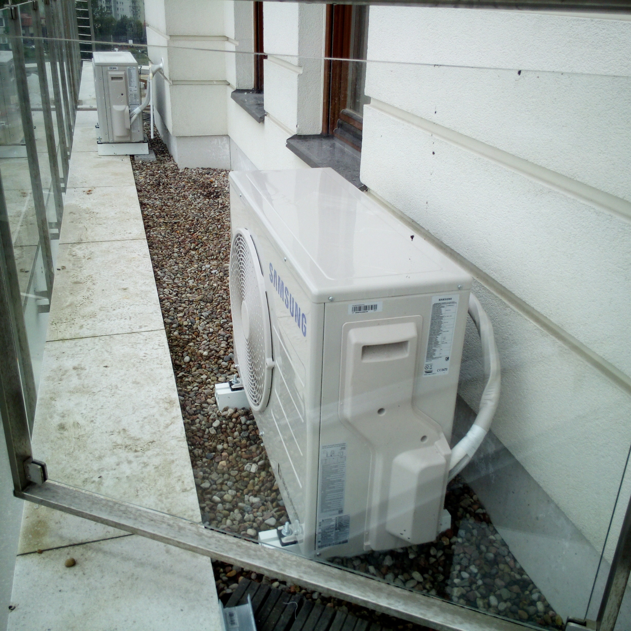 Zamontowany agregat klimatyzacji Samsung na zewnątrz budynku, widoczny przez szklaną balustradę. Tło stanowią małe kamienie i fragment elewacji.
