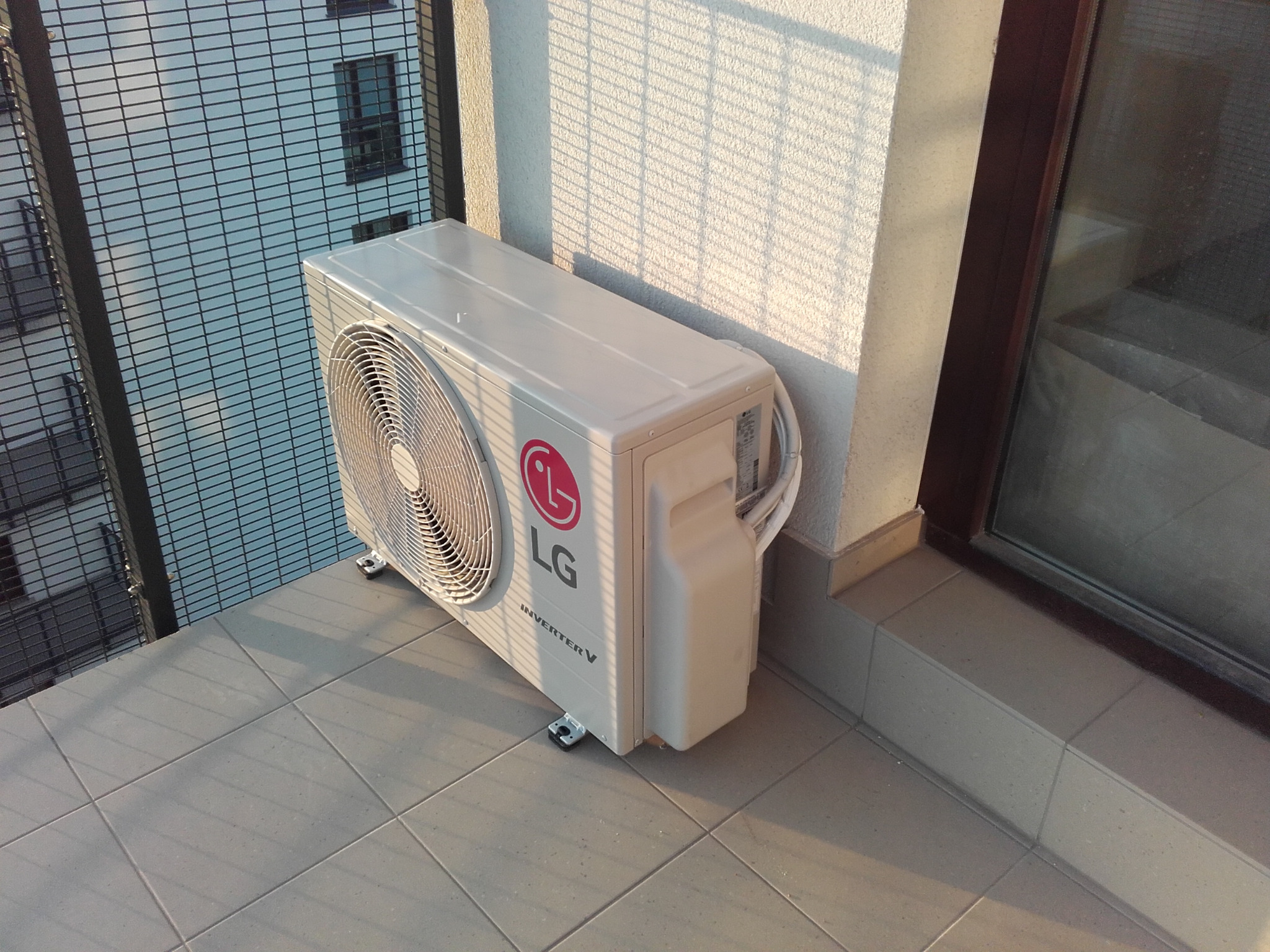 Biały klimatyzator LG Inverter V na balkonie w bloku, oświetlony popołudniowym słońcem, z widoczną elewacją budynku w tle. Montaż klimatyzacji.