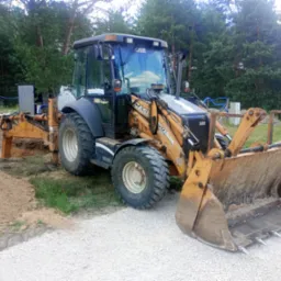 Żółto-szara koparko-ładowarka Case 580 Super L na żwirowej drodze, z wykopem i stertą ziemi w tle, otoczona zielenią i drzewami. Widoczne ślady użytkowania.