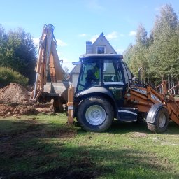 Q1 Dawid Kułan - Pomarańczowa koparko-ładowarka Case 580 Super R w trakcie prac ziemnych, z widocznym wykopem i stertą ziemi na tle domu i drzew.