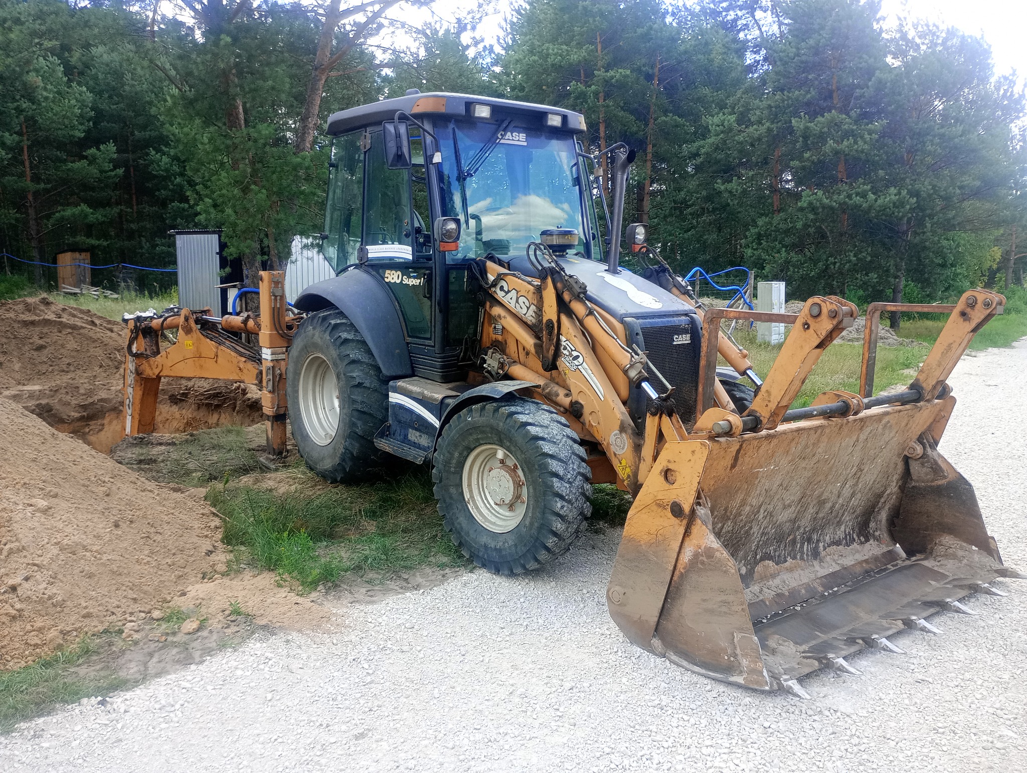 Żółto-szara koparko-ładowarka Case 580 Super L na żwirowej drodze, z wykopem i stertą ziemi w tle, otoczona zielenią i drzewami. Widoczne ślady użytkowania.